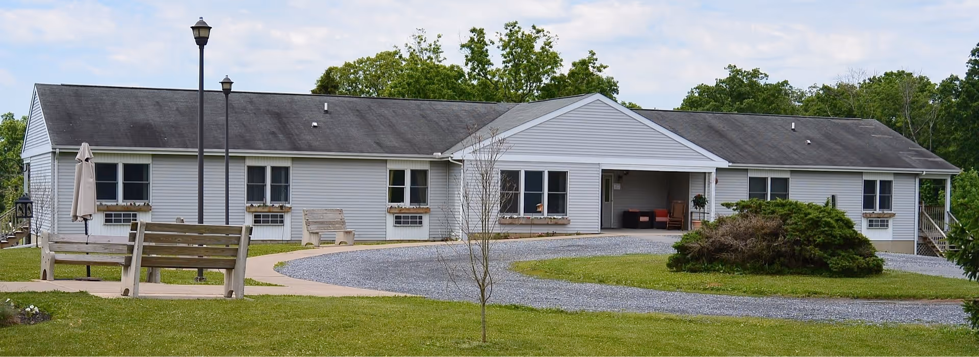 Single-story building with white siding and a dark roof, surrounded by green grass and trees. There are benches, a lamp post, and a small paved circular driveway in front of the building.