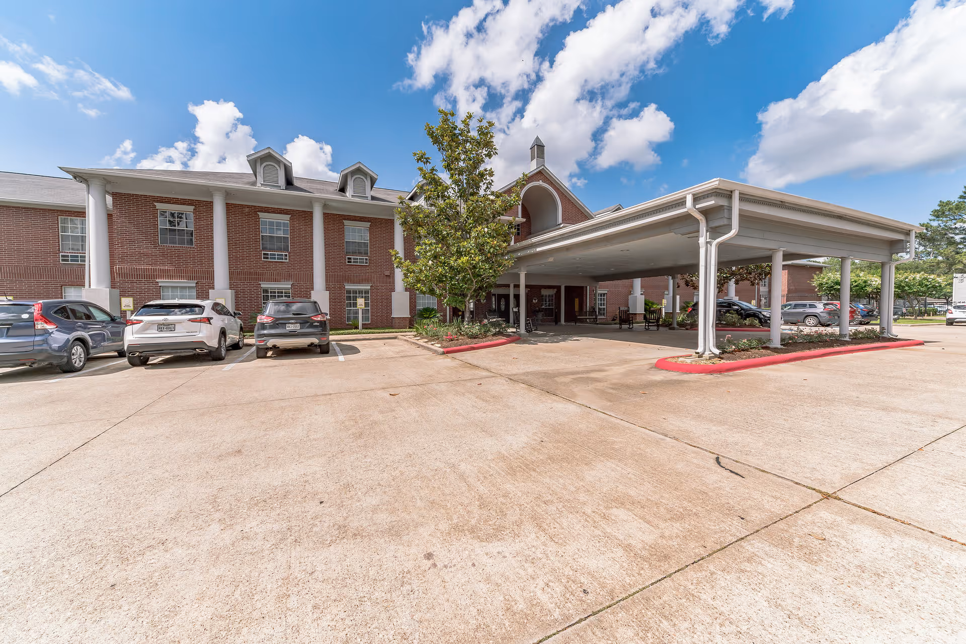Exterior view of Collier Park facility showing a brick building with white columns and a covered entrance area. Several cars are parked in front of the building under a blue sky with scattered clouds.
