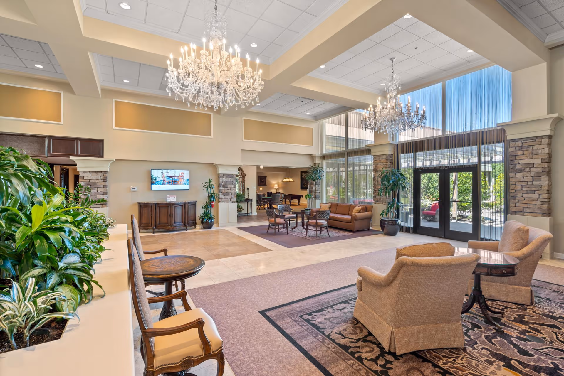 Spacious, well-lit senior living facility lobby with chandeliers, seating areas, plants, and a large glass entrance.