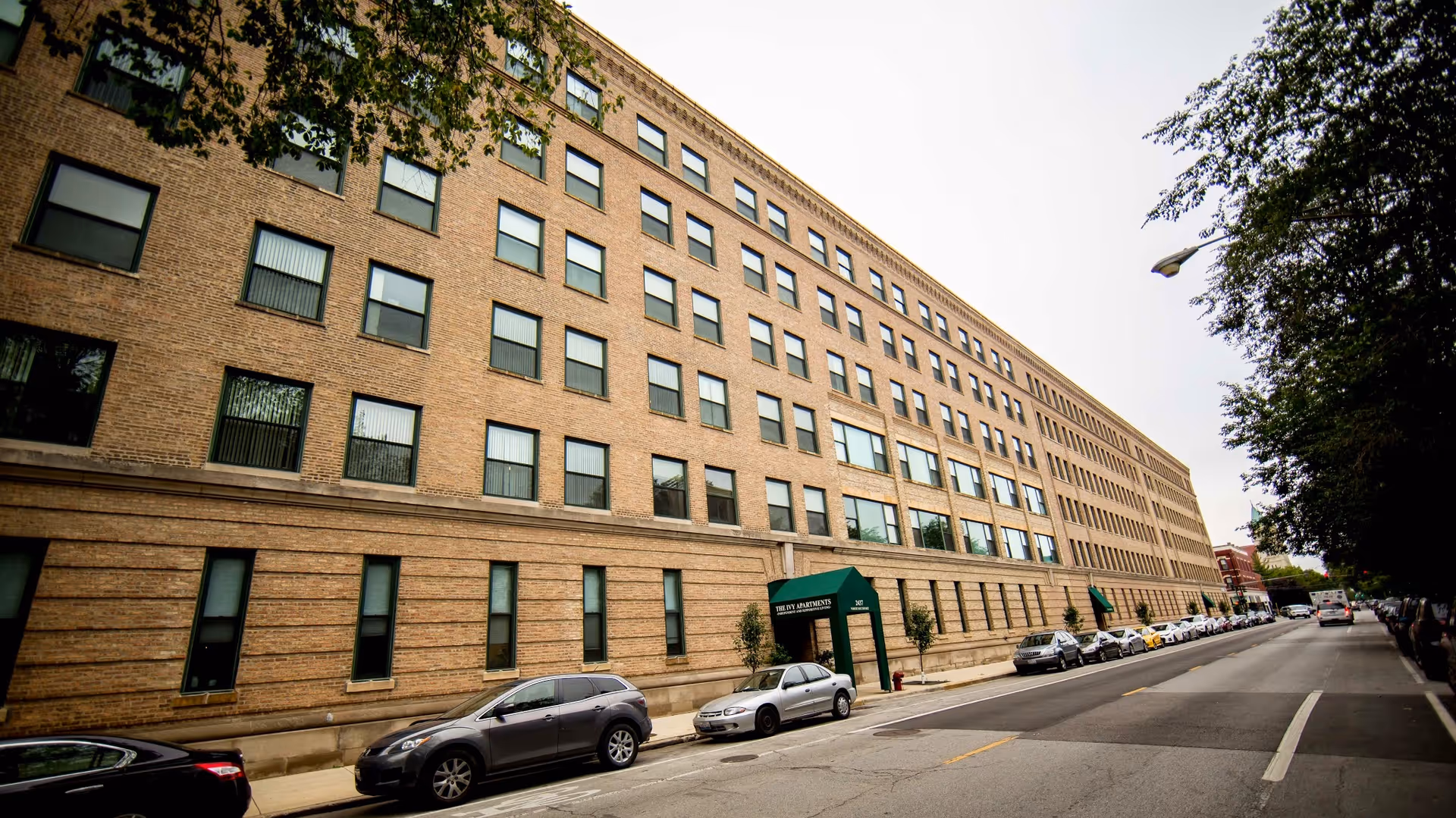 Wide exterior view of a large multi-story brick apartment building with many windows, a green awning entrance, and cars parked along the street in front. Trees line the sidewalk on the right side of the image.