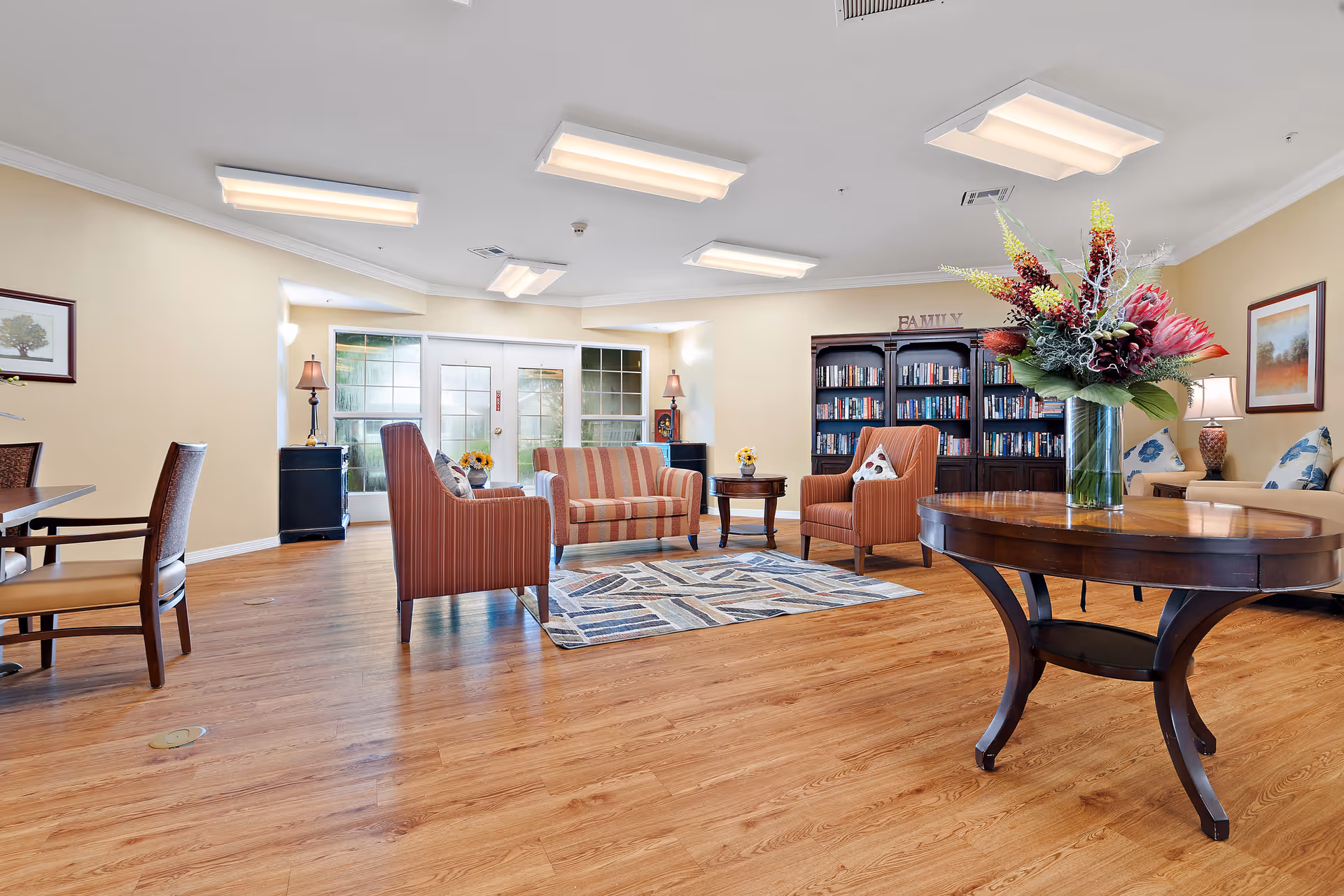 A bright and spacious living room area with wooden flooring, featuring a round wooden table with a large floral arrangement, several upholstered chairs and sofas arranged around a patterned rug, a bookshelf filled with books, framed artwork on the walls, and large windows allowing natural light to enter.