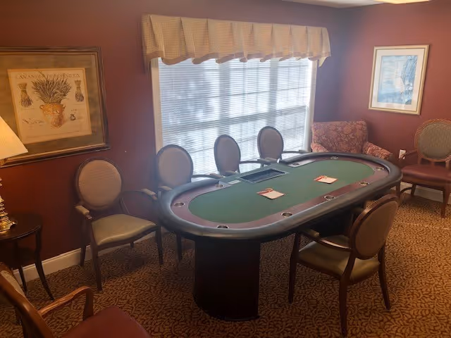 A room with a green felt poker table surrounded by six chairs. The room has maroon walls, a window with blinds and a beige valance, a framed picture on the left wall, a framed artwork on the right wall, a patterned carpet, and a table lamp on a small side table.