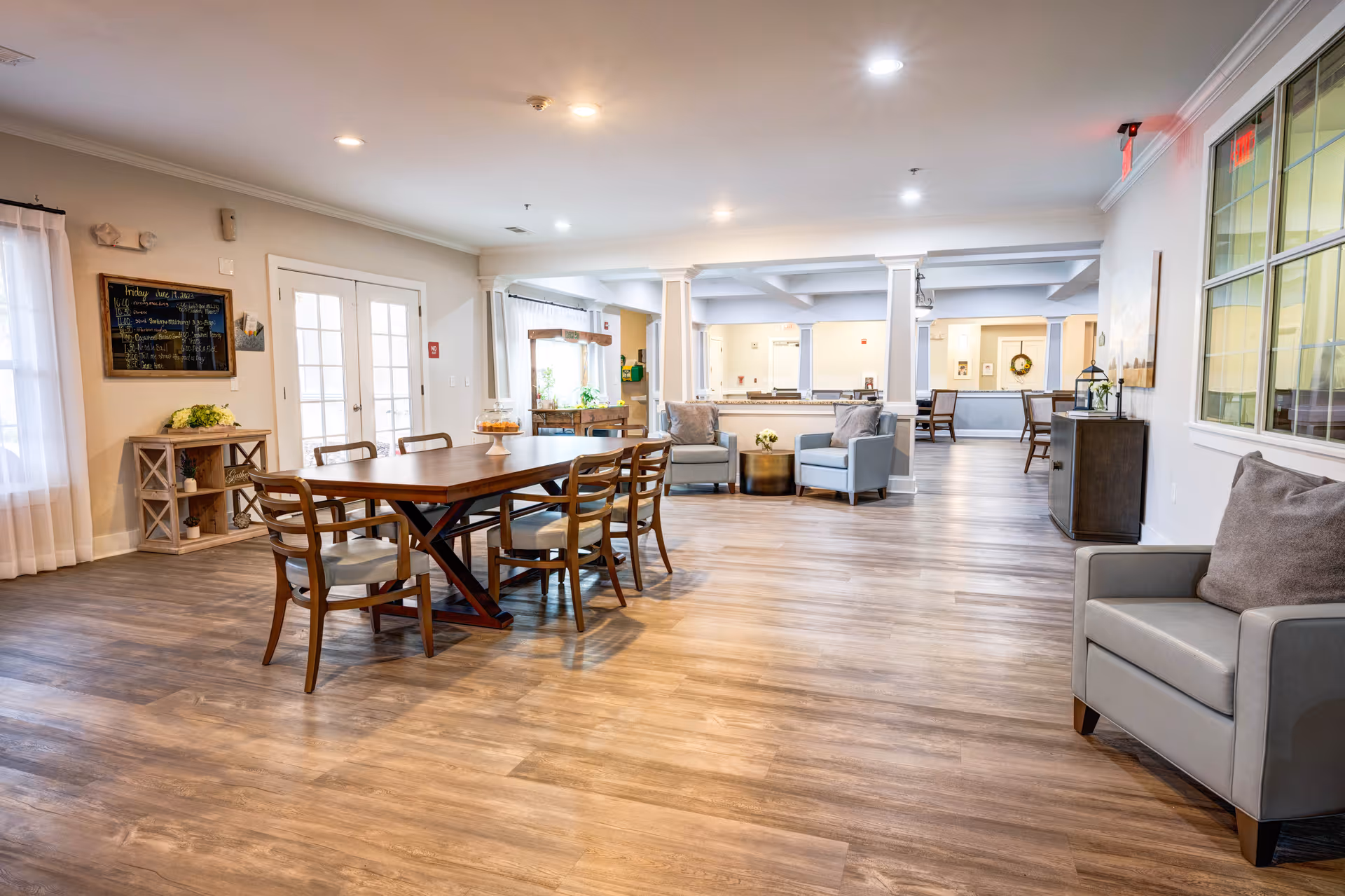A spacious and well-lit common area in an assisted living facility featuring a wooden dining table with six chairs, several comfortable armchairs, and a small side table with a flower arrangement. The room has wood flooring, white walls, large windows with sheer curtains, and ceiling lights. In the background, there is a reception desk and additional seating areas.
