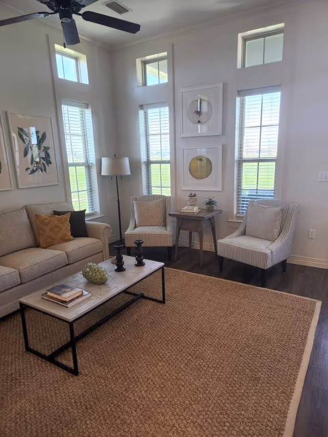 Bright living room with a sofa, two striped armchairs, a coffee table on a woven rug, tall windows, and wall art.