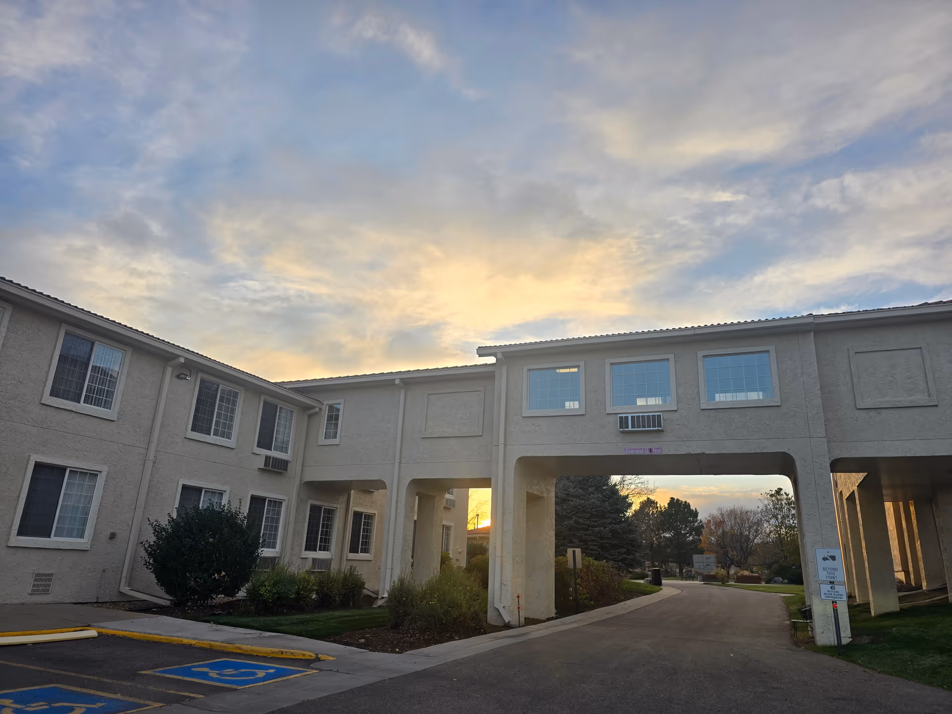 Two-story beige senior living building with an archway driveway entrance, nearby handicap parking spaces, and a sunset sky.