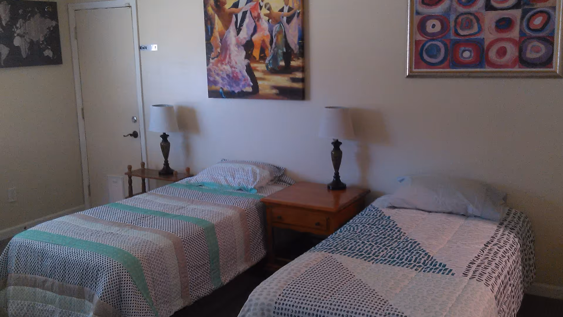 A bedroom with two single beds separated by a wooden nightstand with a lamp on it. Each bed has a different patterned bedspread and a pillow. There are two framed paintings on the wall above the beds, and a closed door is visible in the background.