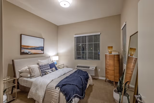 A cozy bedroom with a neatly made bed featuring white and blue bedding. There is a nightstand with a lamp on the left side of the bed, a window with blinds above a wall-mounted air conditioning unit, a tall wooden dresser with a decorative lamp and books, and a large floor mirror leaning against the wall. A potted plant is placed near the mirror, and a framed abstract artwork hangs above the bed.