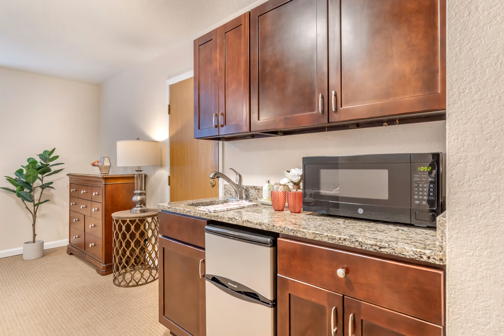 A small kitchenette with granite countertop, dark wood cabinets, a microwave, mini fridge, sink, and a dresser with a lamp and potted plant in the background.