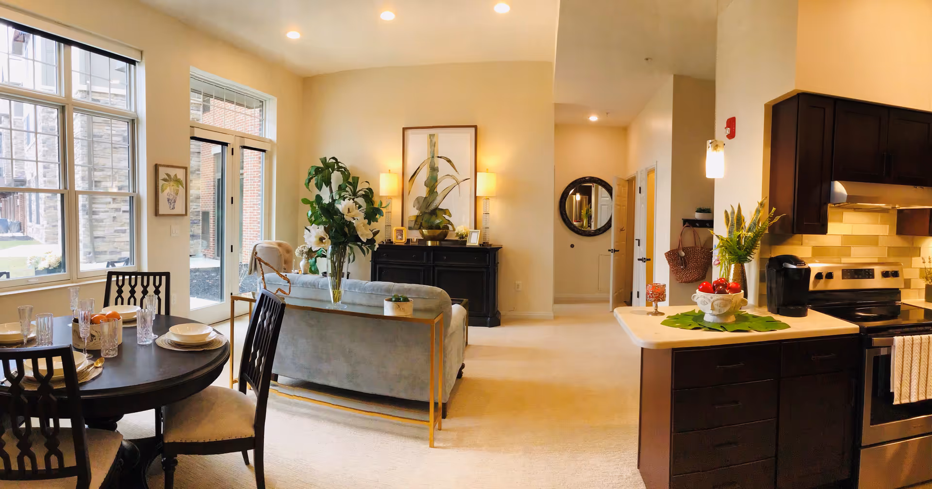 Open-concept living area with a sofa, dining table, kitchen island, and large windows in a well-lit senior living apartment.