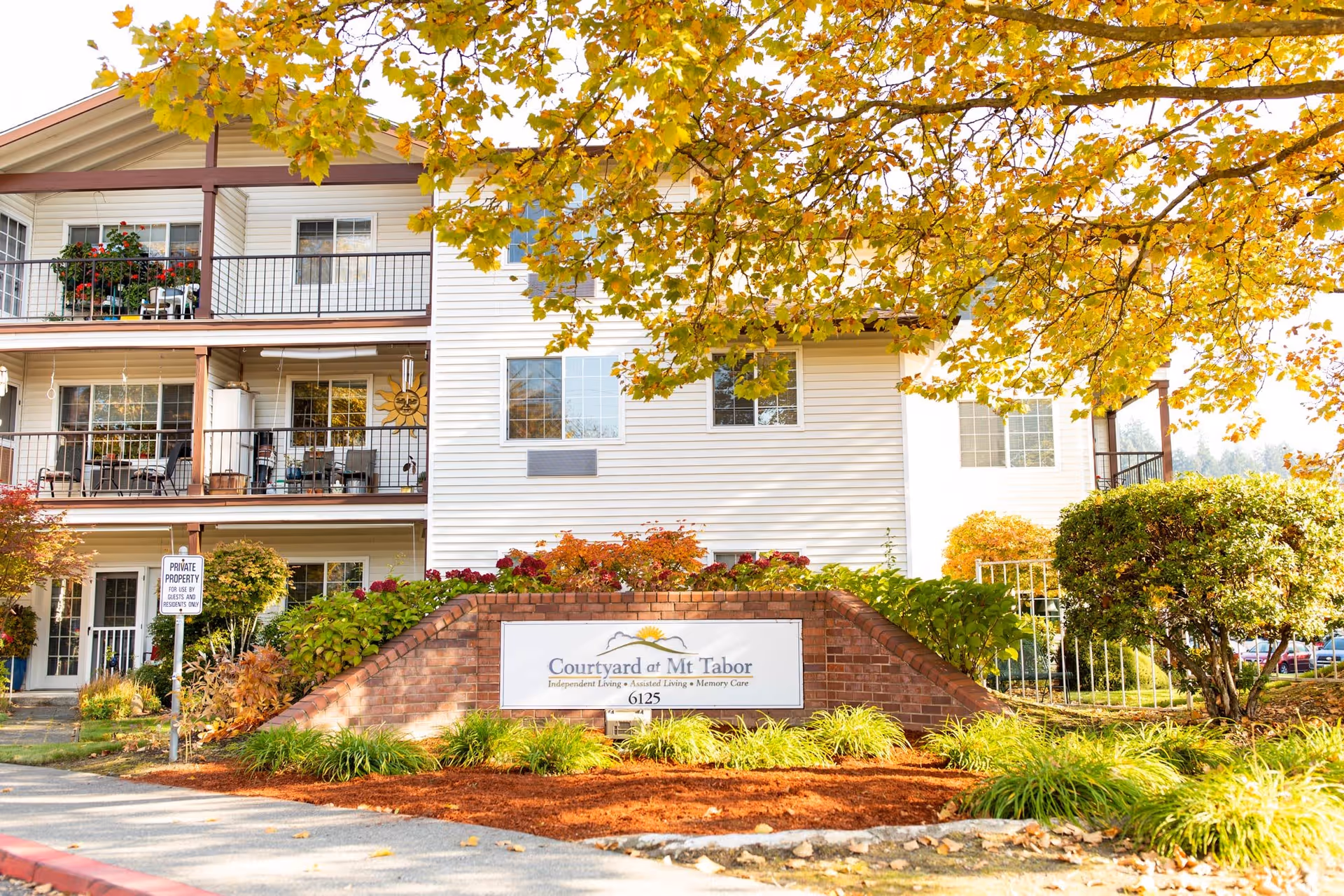 Exterior view of Courtyard at Mt. Tabor senior living facility with a brick sign in front displaying the facility name and services. The building is white with balconies and surrounded by autumn foliage and landscaping.