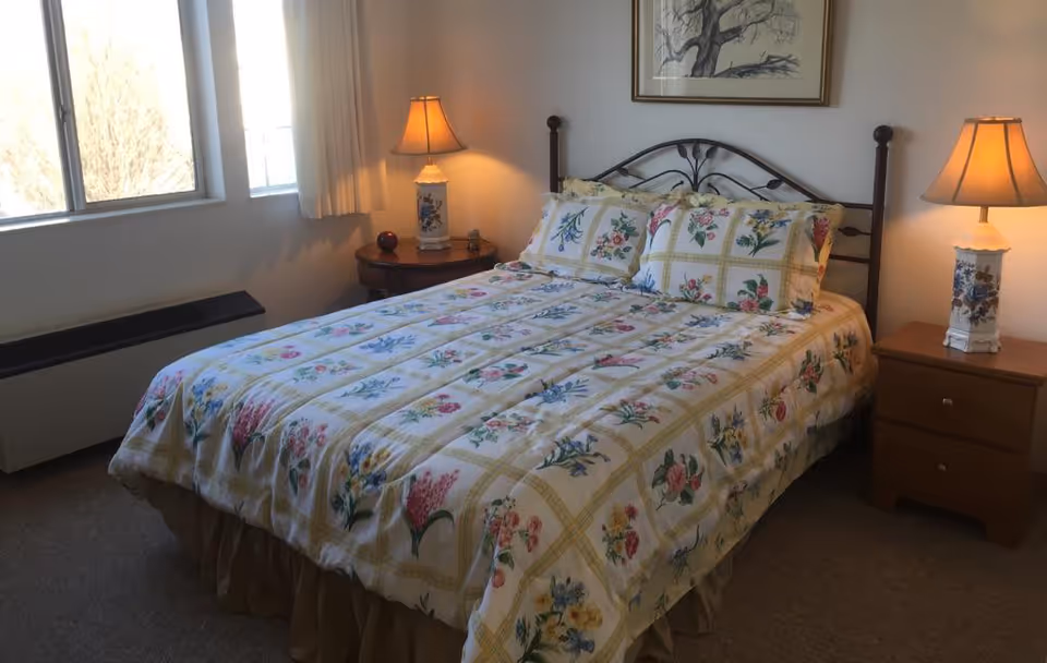 A bedroom with a bed covered in a floral patterned bedspread and matching pillows. There are two bedside tables, each with a lamp. A window with white curtains is on the left side, and a framed picture hangs above the bed.