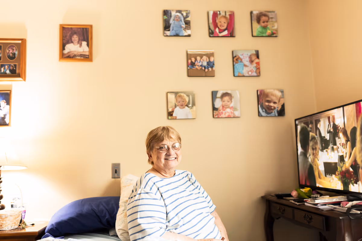 A smiling person sits on a bed in a decorated bedroom with framed photos on the wall and a television on a dresser.