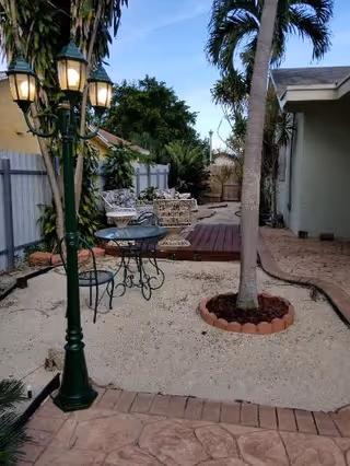 Outdoor patio area with a tall lamp post, a small round metal table with two matching chairs, a palm tree planted in a circular brick border, and a wooden deck with white wicker furniture surrounded by plants and a fence.