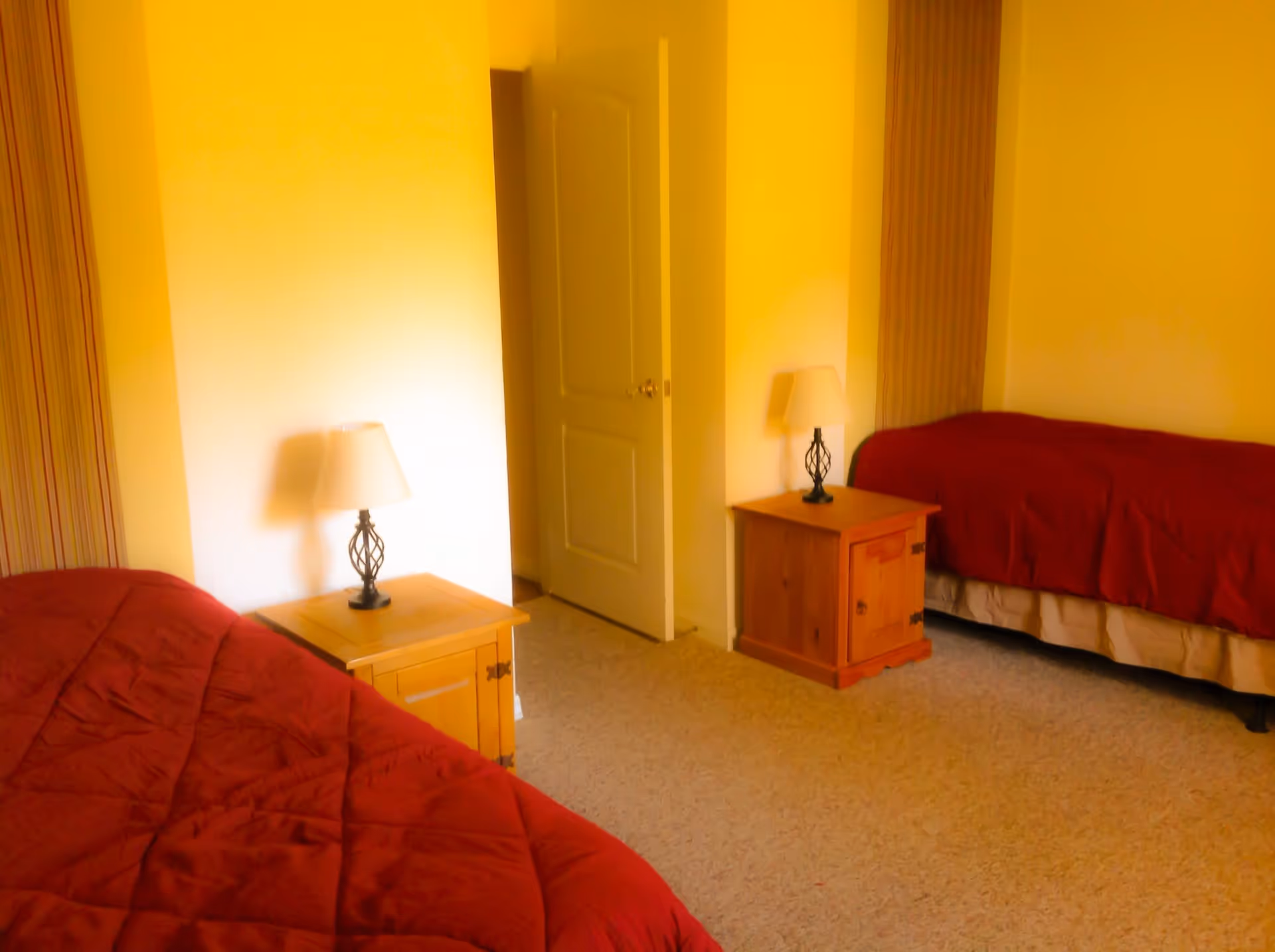 A warm-lit bedroom with two twin beds dressed in red quilts, wooden nightstands with lamps, and a door in the center background.