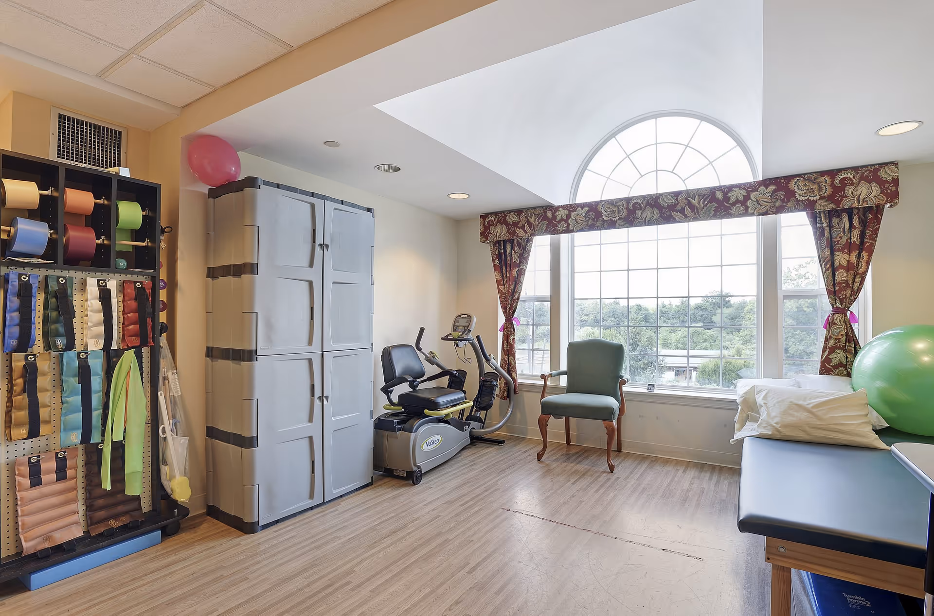 Bright therapy/activity room with exercise equipment, storage cabinets, a treatment table, and a large arched window.