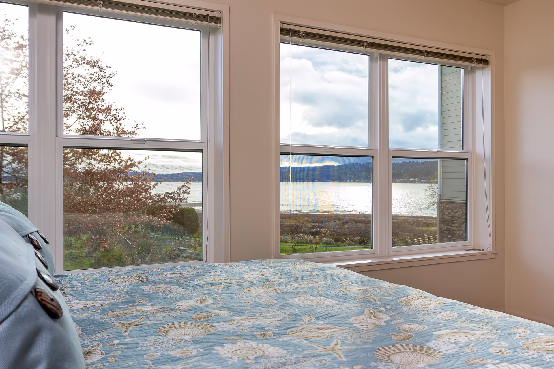 A bedroom with a bed covered in a blue quilt featuring seashell patterns, positioned next to large windows that offer a scenic view of a lake, trees, and hills under a cloudy sky.