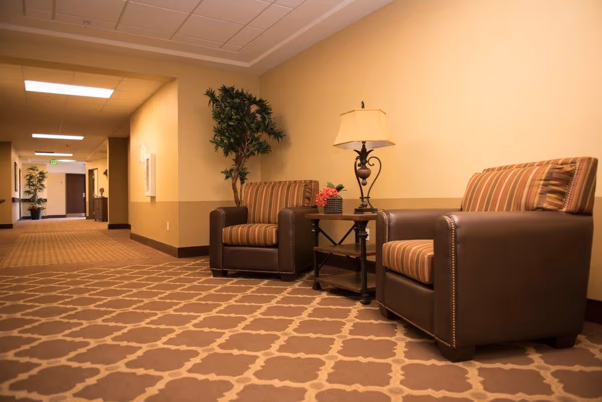 A hallway in a senior living facility with two brown leather armchairs featuring striped cushions, a small wooden side table with a decorative lamp and a small plant, and a tall potted plant against a beige wall. The hallway has patterned carpet and overhead fluorescent lighting.