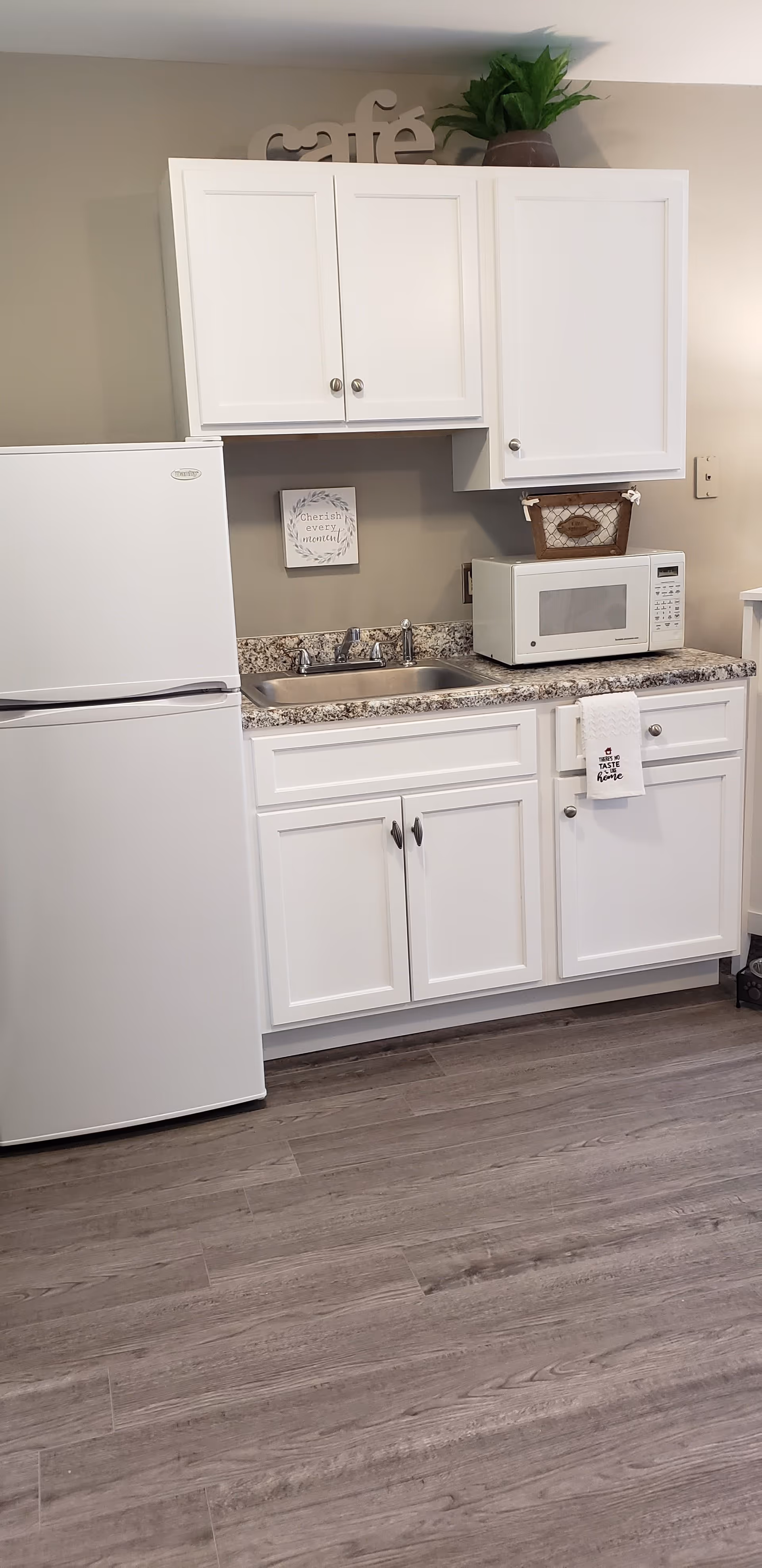A small kitchen area with white cabinets, a white refrigerator, a microwave on the countertop, a sink with a faucet, and a granite countertop. Above the cabinets is a decorative sign that says 'café' and a potted plant. A small framed sign on the wall reads 'Cherish every moment'. A white towel hangs from a cabinet handle with the text 'There's no taste like home'. The floor is a light wood laminate.