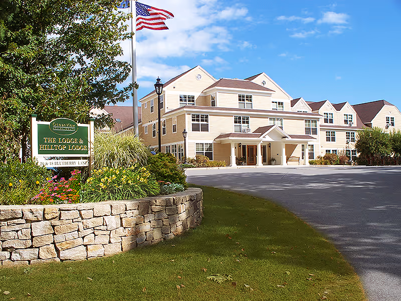 Front exterior of a multi-story beige senior living building with a driveway, landscaped stone planter, entrance canopy, an American flag, and a sign for The Lodge & Hilltop Lodge.
