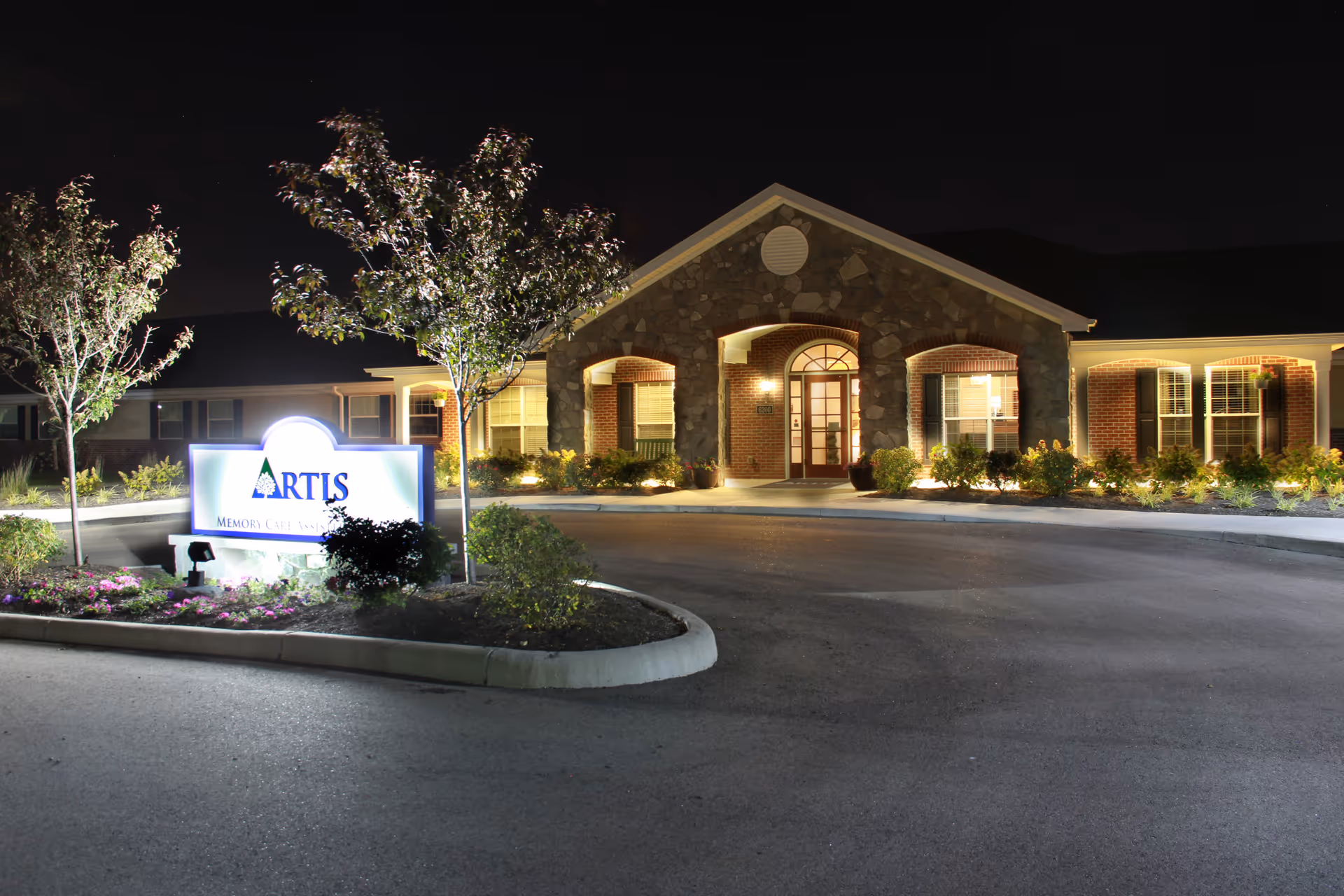 Night view of the front entrance of Artis Senior Living of Mason, featuring a well-lit stone and brick building with arched doorways and windows, surrounded by landscaped bushes and trees. A bright sign with the facility's name is visible near the driveway.