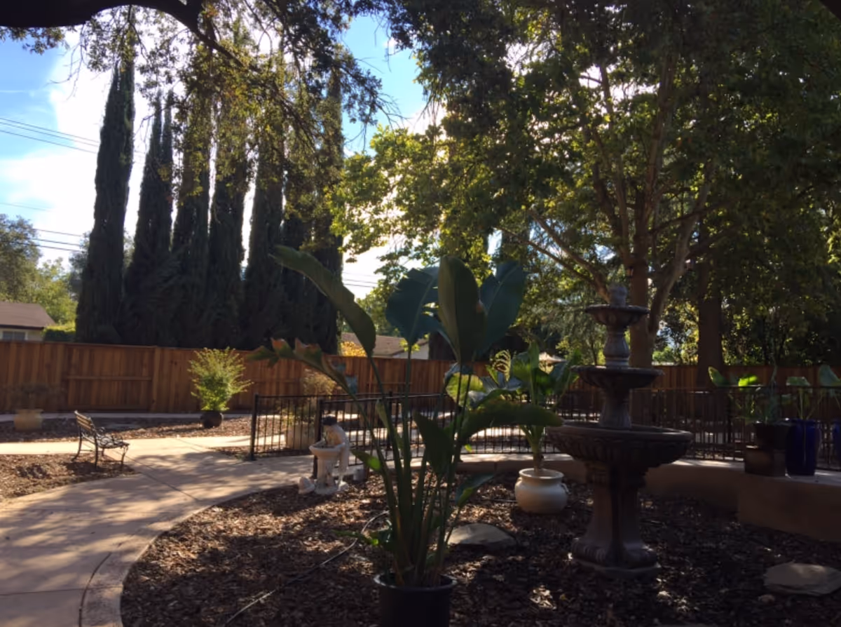 Shaded outdoor courtyard with a multi-tiered stone fountain, potted plants, benches, and a curved walkway surrounded by trees and a wooden fence.