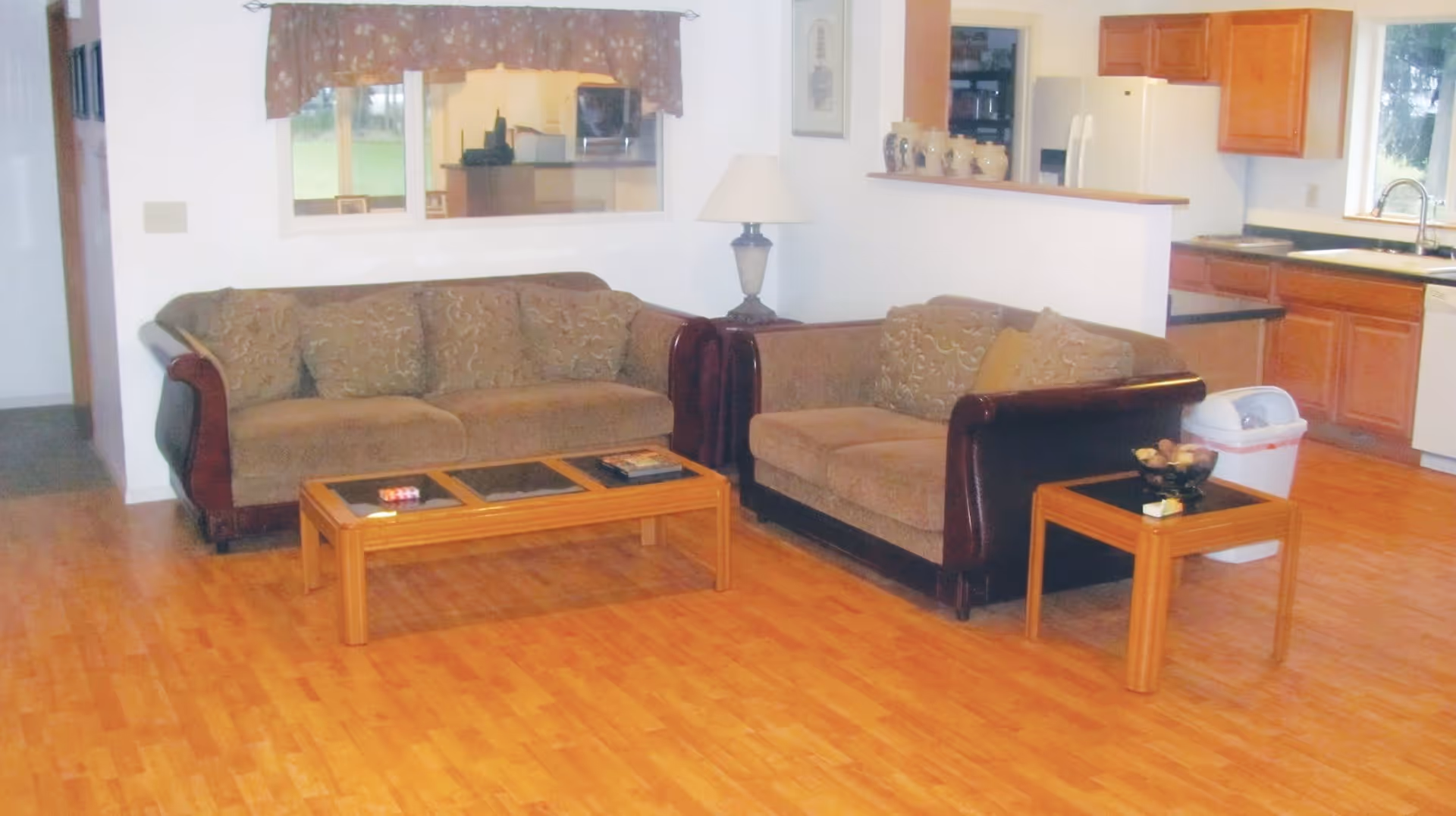 A living room area with two brown cushioned sofas with dark wooden arms, a wooden coffee table with items on it, and a wooden side table with a decorative bowl. Behind the sofas is a half wall separating the living room from a kitchen area with wooden cabinets, a sink, and a white trash bin. The floor is a light wood laminate, and there is a window with a valance above one sofa.