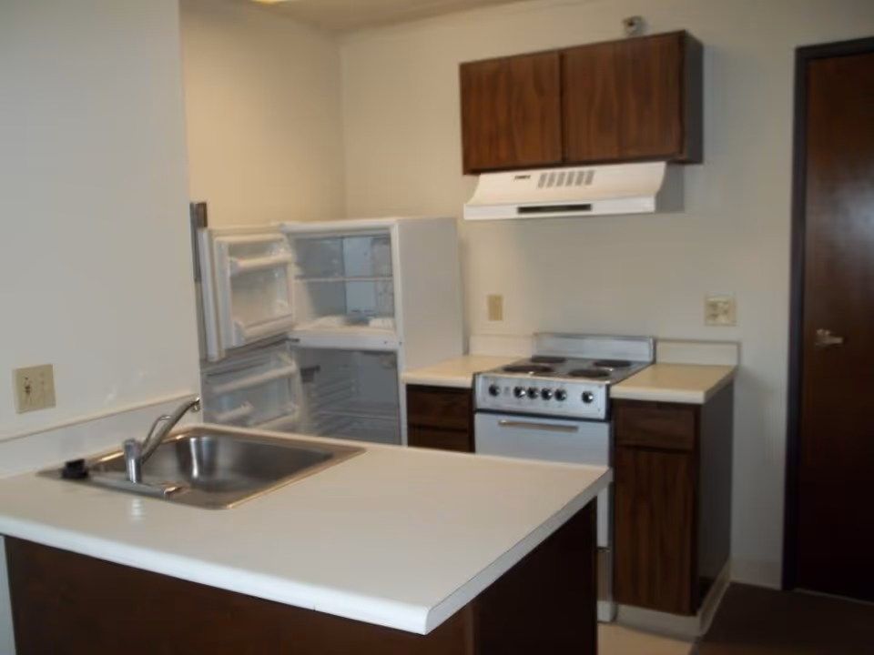 Small kitchen area with a white countertop island featuring a stainless steel sink and faucet. In the background, there is an open white refrigerator, a stove with four burners, a range hood, and wooden cabinets mounted on the wall and below the counter. The walls are light-colored, and there is a closed dark wooden door on the right side.