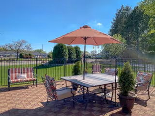 Outdoor patio area with a square table and four cushioned chairs under a large beige umbrella. Two cushioned benches are placed along the black metal fence surrounding the patio. Green grass, trees, and a clear blue sky are visible in the background.