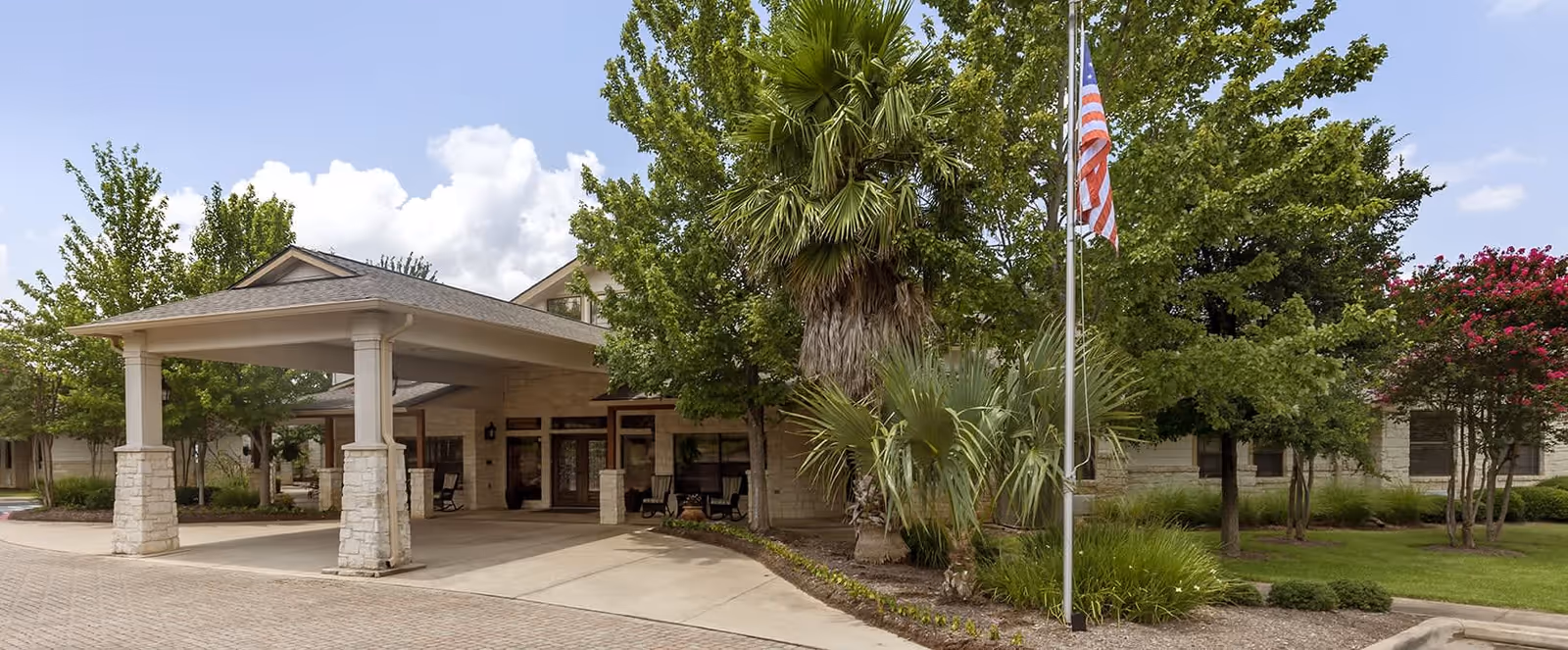 Front exterior view of Brookdale Lohmans Crossing facility with a covered entrance, stone pillars, trees, and an American flag on a flagpole.