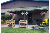 Outdoor patio area with a green and white striped awning attached to a building. The patio has hanging flower baskets, two wooden planters with flowers, a bench, and a small table. The area is surrounded by green grass and some landscaping with bushes and yellow flowers.