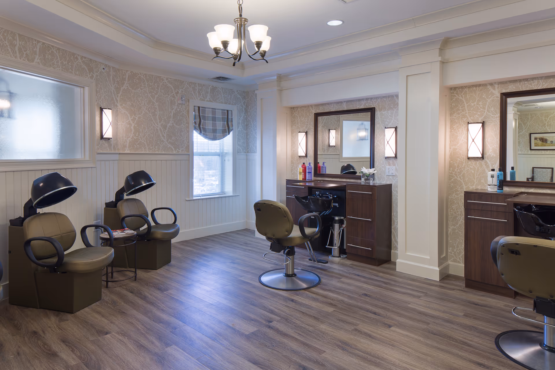 Interior of a senior living facility hair salon with two hair drying chairs on the left and two styling stations with mirrors and chairs on the right. The room has wood flooring, patterned wallpaper, a window with a plaid valance, and a chandelier light fixture on the ceiling.