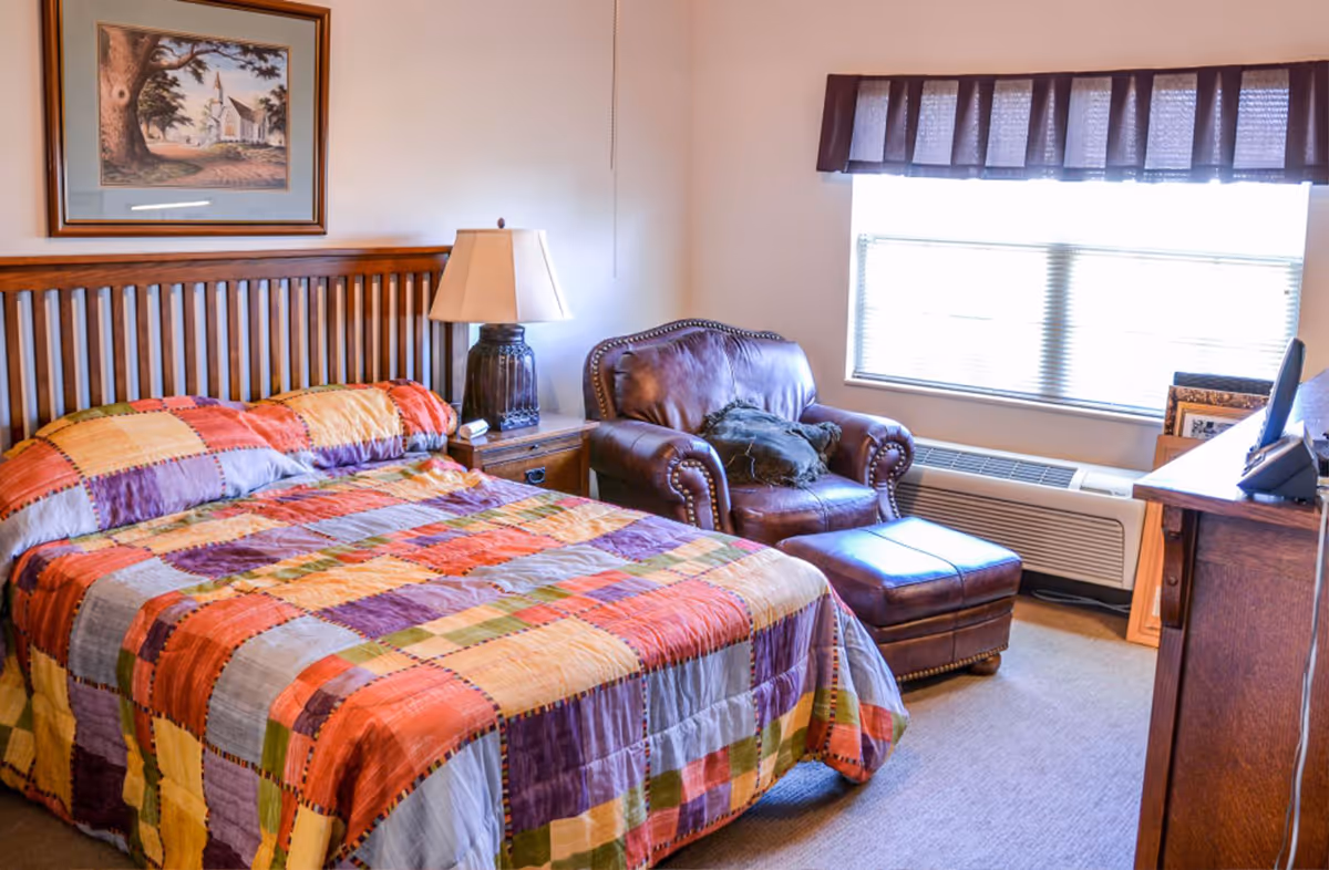 A cozy bedroom with a wooden headboard bed covered in a colorful patchwork quilt. Next to the bed is a wooden nightstand with a lamp. There is a brown leather armchair with an ottoman near a window with blinds and a valance. A framed picture hangs on the wall above the bed, and a wooden dresser with a TV on top is positioned opposite the bed.