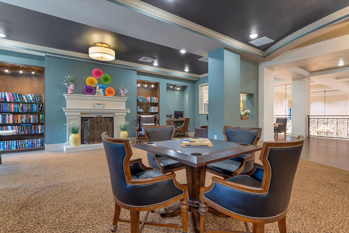 A cozy senior living common area with a square wooden game table surrounded by four leather chairs. In the background, there is a white fireplace decorated with colorful paper fans and vases with flowers. Built-in bookshelves filled with books flank the fireplace. The room has teal walls, a dark ceiling with recessed lighting, and a carpeted floor with a leopard print pattern. There is also a computer desk with a chair in a small alcove.