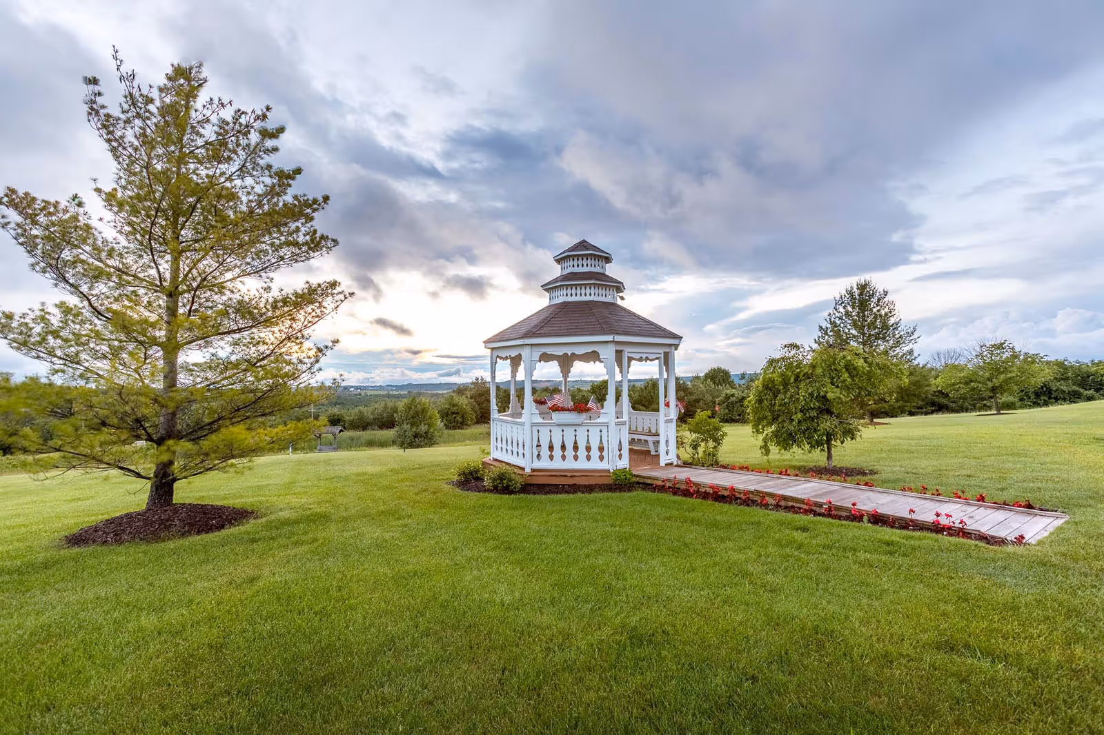 A white wooden gazebo with a brown roof sits on a well-maintained green lawn surrounded by trees and flower beds under a cloudy sky.