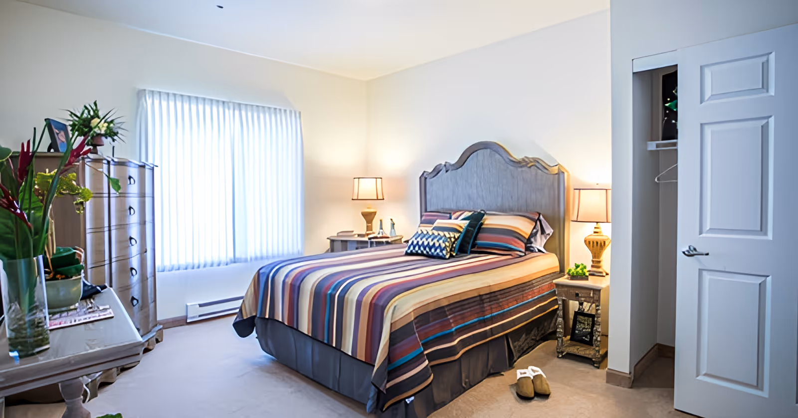 A bright bedroom with a large window covered by sheer white curtains. The room features a bed with a striped multicolored bedspread and several pillows, two bedside tables each with a lamp, a dresser with a framed photo and plants, and an open closet door revealing hangers inside. There are slippers placed on the carpeted floor near the bed.