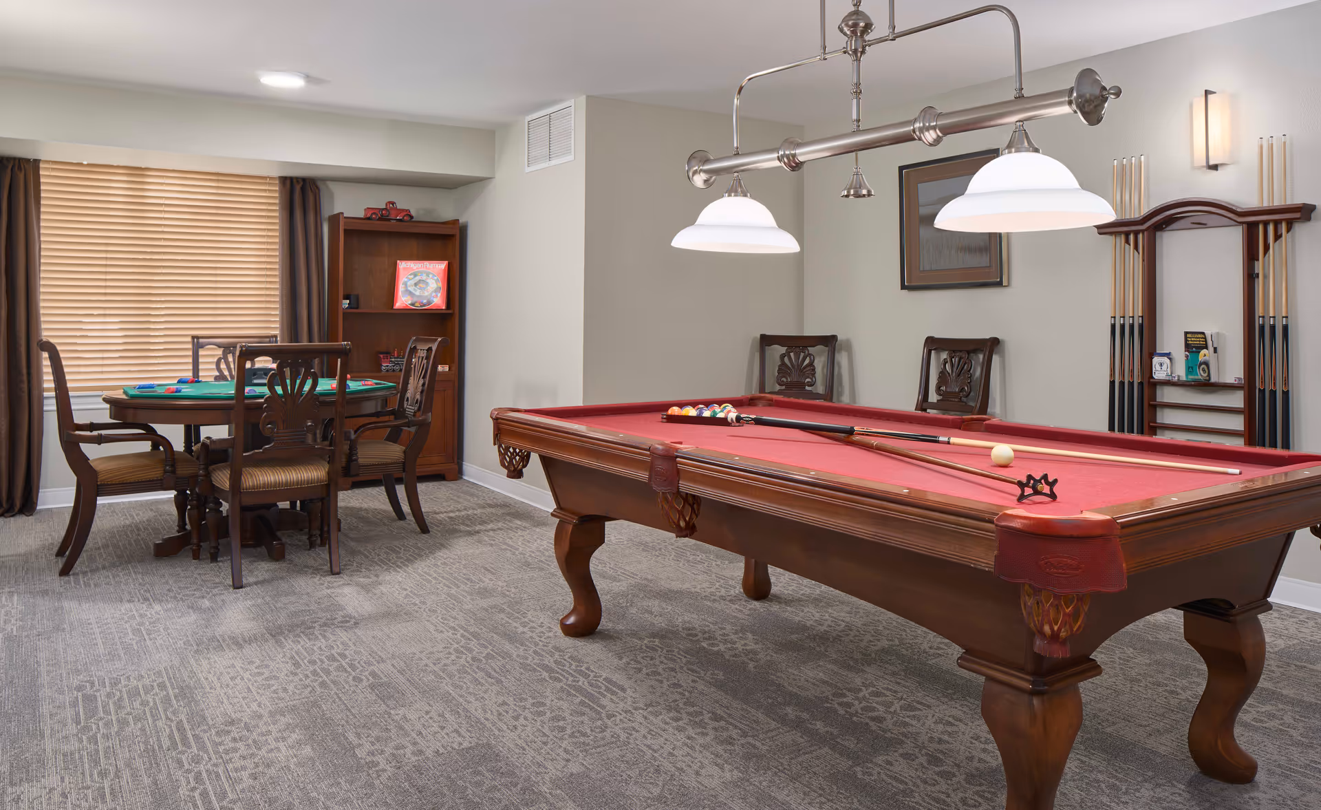 A recreation room featuring a red-felt pool table with cues, overhead lights, a cue rack, and a nearby card table with chairs.