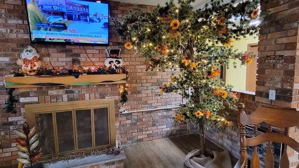 Cozy brick-walled sitting area with a fireplace, wall-mounted TV, decorated mantel and a lit artificial tree with sunflowers.