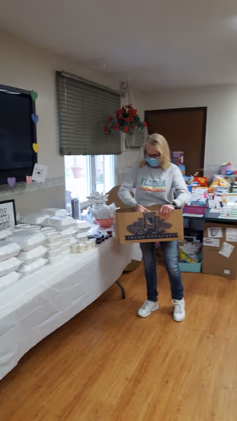 A woman wearing a face mask and a Florida sweatshirt is holding a cardboard box labeled 'Idaho Potatoes' in a room with wooden flooring. To her left, there is a table covered with a white tablecloth holding multiple stacked white takeout containers and small jars. Behind her, there are shelves and tables filled with various supplies and boxes. A window with closed blinds and a hanging plant are visible on the wall.