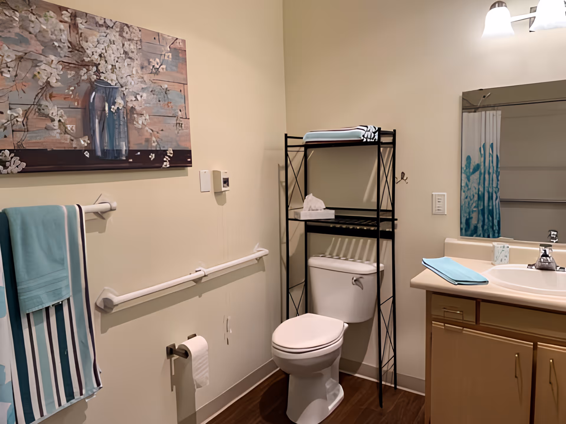 A bathroom with a toilet, a black metal shelving unit above the toilet holding folded towels and tissues, a sink with a countertop and a mirror above it, a towel rack with striped and solid teal towels, a wall-mounted grab bar, and a decorative painting of flowers in a vase on the wall.