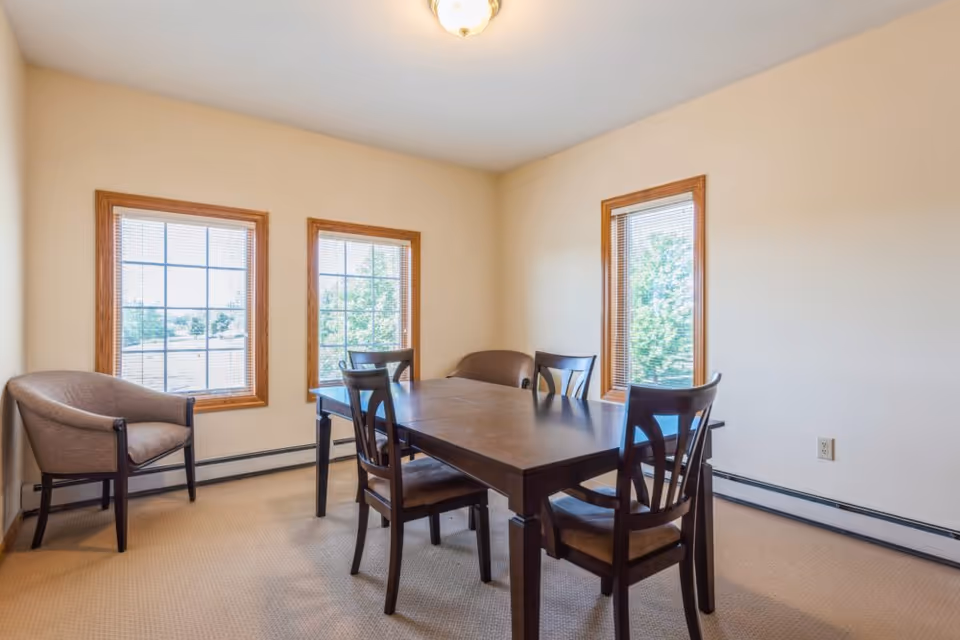 A small dining area with a rectangular wooden table surrounded by four chairs. Two windows with wooden frames let in natural light, and two cushioned armchairs are placed against the walls. The room has beige walls and carpeted flooring.