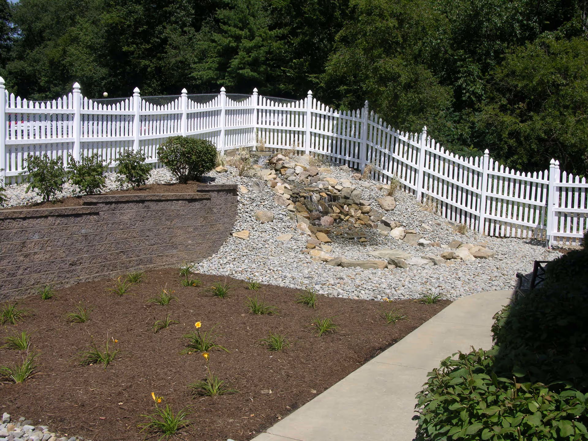 A landscaped outdoor area featuring a white picket fence, a rock-lined dry creek bed, planted mulch beds, and a concrete walkway.