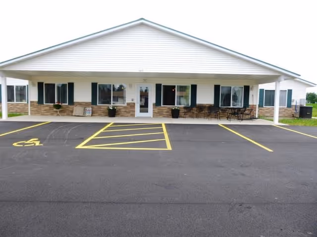 Front exterior view of a single-story assisted living facility building with a covered entrance, several windows, outdoor seating, and a parking lot with marked spaces including a handicapped spot.