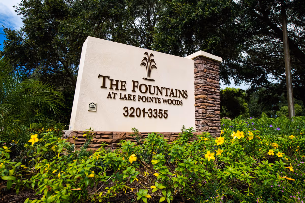 Stone and stucco entrance sign reading "The Fountains at Lake Pointe Woods 3201-3355" surrounded by flowering landscaping and trees.