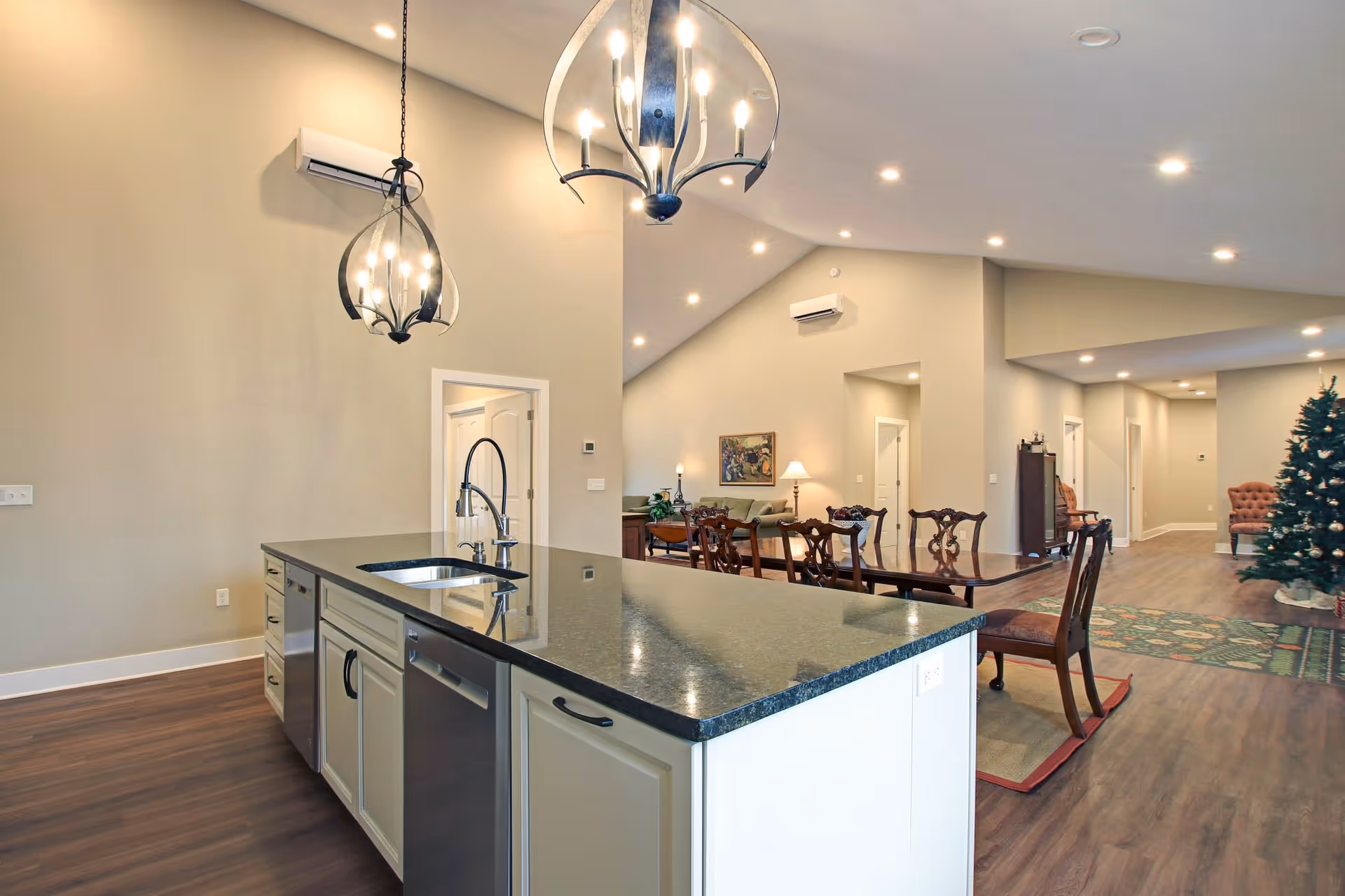 Interior view of a senior living facility showing a modern kitchen island with a black countertop and built-in sink, two hanging pendant lights above, and a dining area with wooden chairs and a table in the background. The space has high ceilings with recessed lighting, light-colored walls, wood flooring, and a decorated Christmas tree near the hallway.