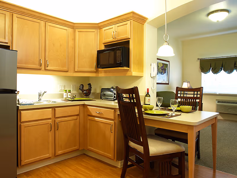 A cozy kitchen area with light wood cabinets, a stainless steel refrigerator, a microwave, a toaster, and a sink. Adjacent to the kitchen is a small dining table set with two chairs, plates, bowls, and glasses. In the background, there is a living area with a window covered by a green valance and an air conditioning unit below it.