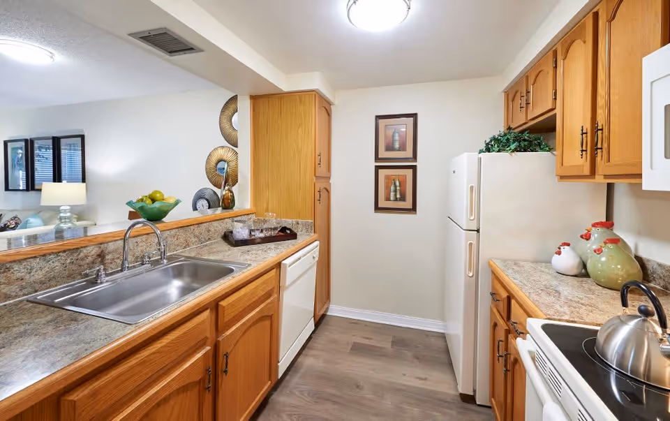 A bright kitchen with wooden cabinets, a double stainless steel sink, a white dishwasher, a white refrigerator, and a white stove with a kettle on it. The countertops are a speckled beige color. There are two framed pictures on the wall and decorative items including a bowl of fruit and ceramic chickens. The kitchen opens to a living area with a lamp and wall decorations visible.