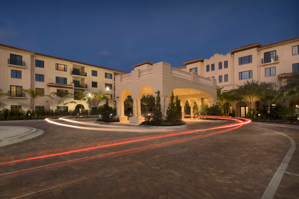 The illuminated porte-cochere entrance of a multi-story senior living building at dusk with car light trails in the driveway.