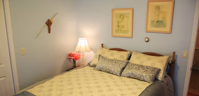 A cozy bedroom with a double bed covered in a patterned quilt and decorative pillows. There is a bedside table with a lamp and a vase of pink flowers. The walls are painted light blue and decorated with two framed pictures of children and a wooden cross with a feather hanging on the left wall.