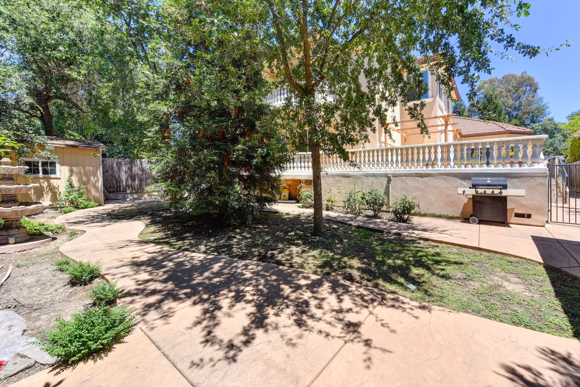 Outdoor garden area with a paved walkway, trees, shrubs, a small shed, a multi-tiered stone fountain, and a barbecue grill next to a beige building with a white balustrade under a clear blue sky.