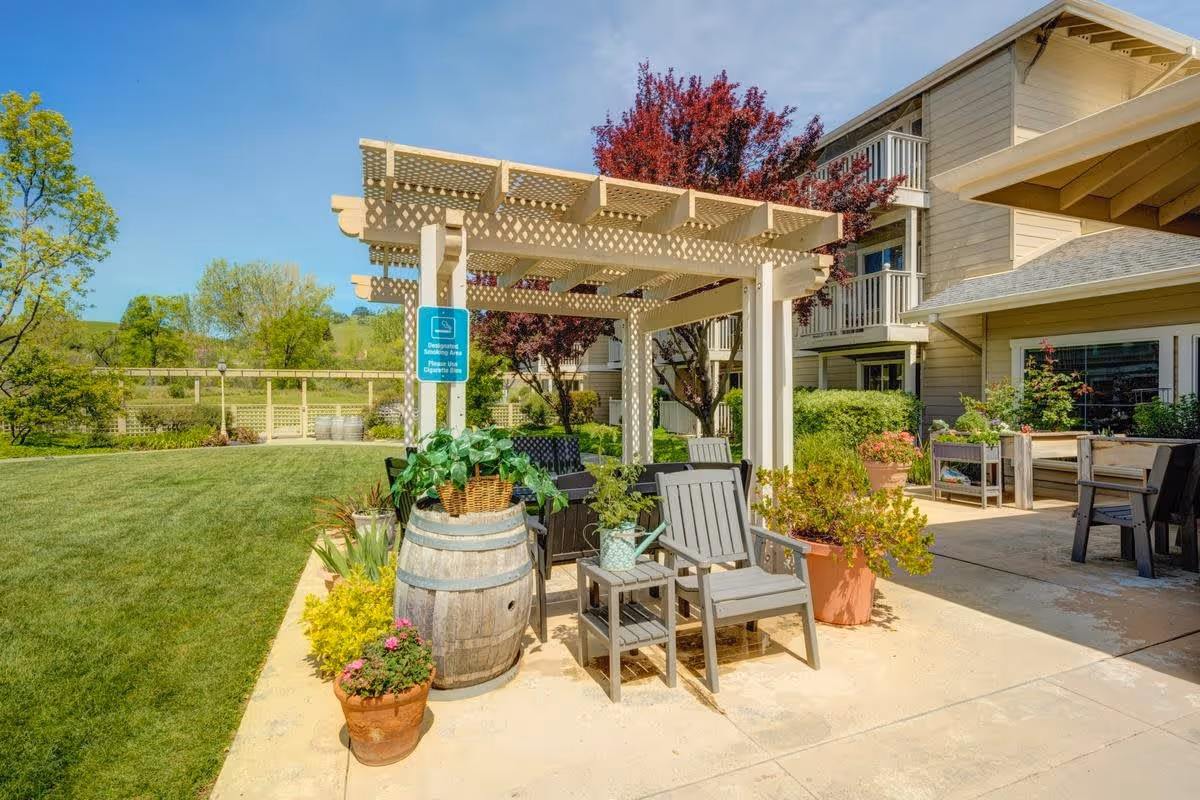 Outdoor patio area at The Village at Rancho Solano featuring a pergola with seating including wooden chairs and a small table. The area is decorated with various potted plants and flowers. In the background, there is a beige multi-story building with balconies and lush green trees and grass under a clear blue sky.